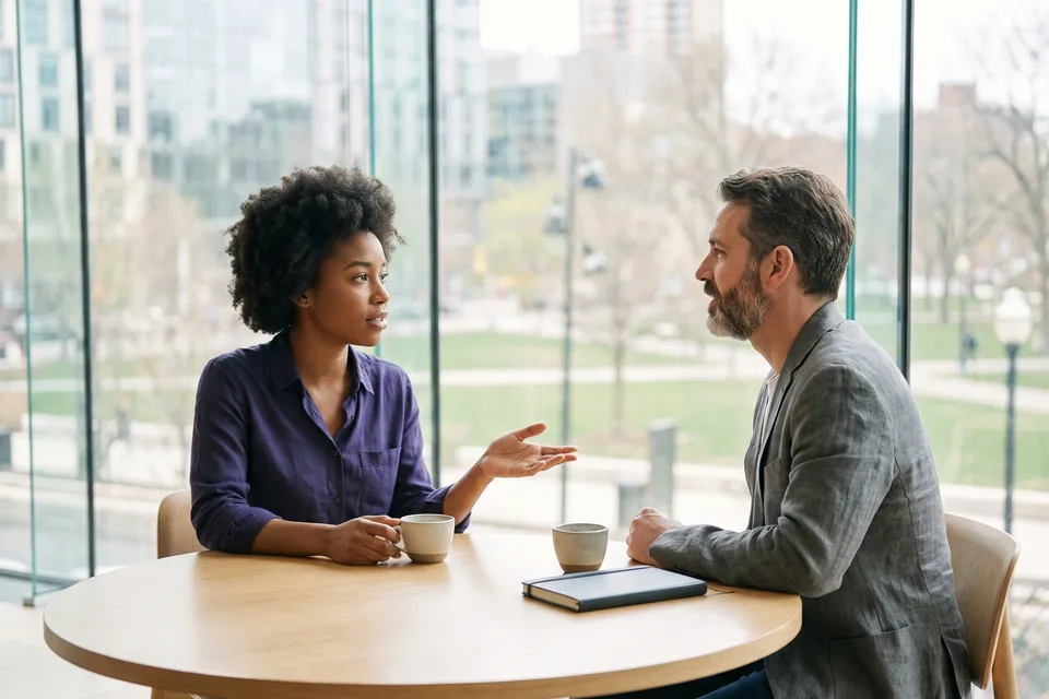 Two colleagues talking at a table in a bright meeting room, she speaks and he listens