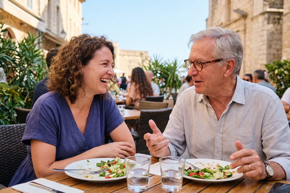 A woman and an older man laughing together over lunch on a sunny restaurant terrace